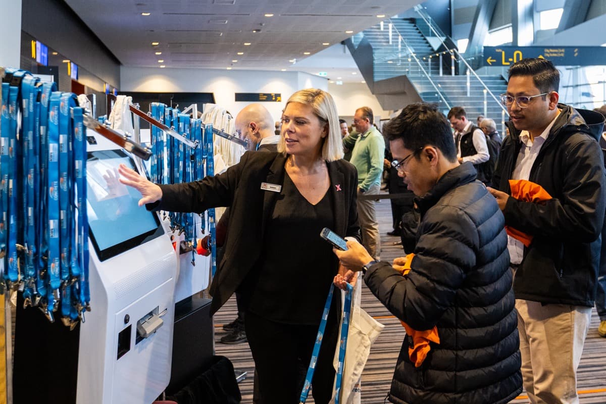 A staff member helps delegates with their phones at a row of check-in kiosks and lanyards